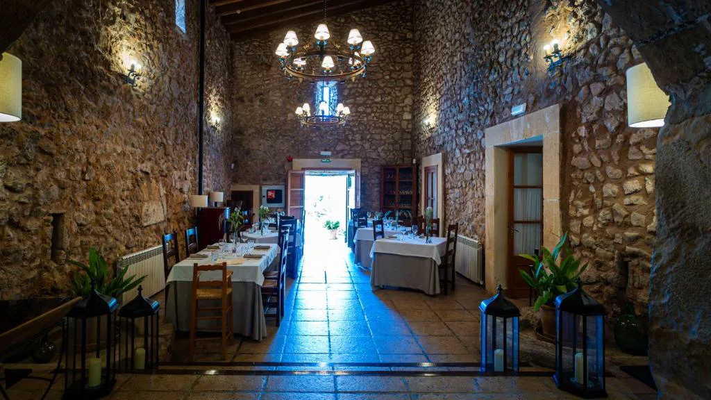 Comedor con paredes de piedra vista, arañas de cristal y mesas vestidas de blanco, Agroturisme Son Cardaix