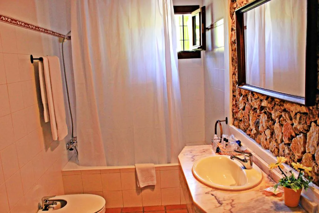 Baño con pared de piedra vista y cortina blanca junto a la bañera, Agroturismo Can Fuster