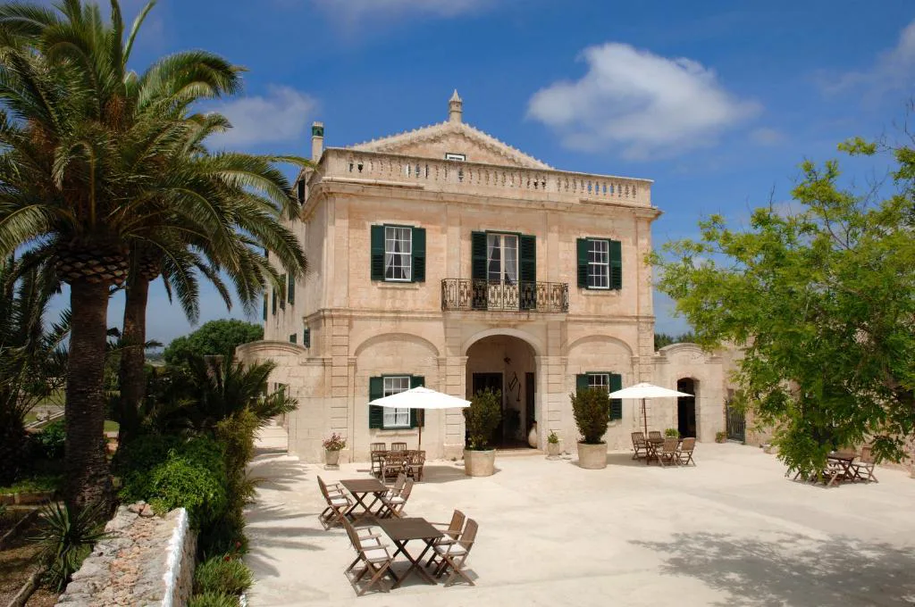 Fachada de piedra dorada del siglo XVIII con palmeras y terraza de mesas de madera, Alcaufar Vell, Sant Lluís, Menorca