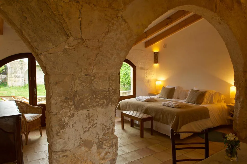 Dormitorio bajo arco de piedra vista con vigas de madera y cama con ropa de cama en tonos verdes, Alcaufar Vell