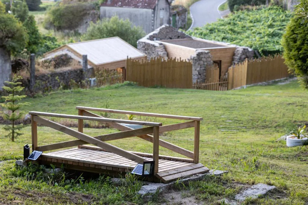Vista de las cabañas de piedra y madera de As Alvarizas entre jardines, con puente decorativo en primer plano