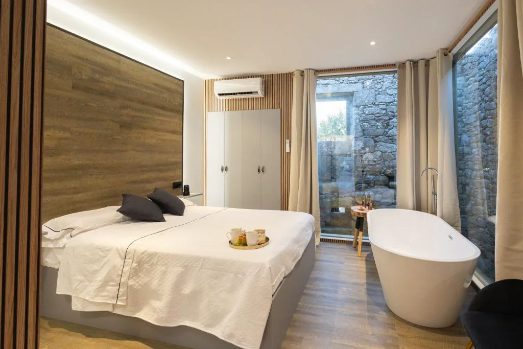 Bañera exenta junto a ventanal de piedra vista y bandeja de café en la cama, As Alvarizas