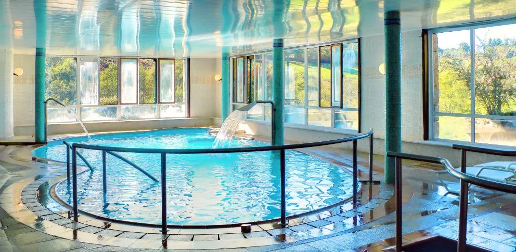 Piscina termal interior con chorros de agua activos bajo cristaleras al bosque, Hotel Balneario Río Pambre