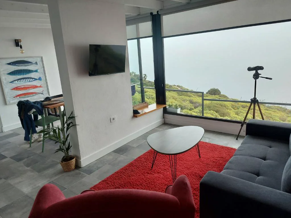 Salón con sofás rojo y azul frente a un ventanal con vistas al mar y un telescopio, Bungalowmirador