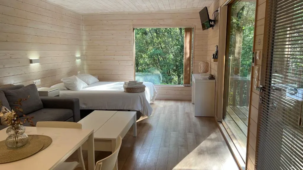 Dormitorio de madera clara con cama doble y vistas al bosque a través del ventanal, en Cabanas das Chousas