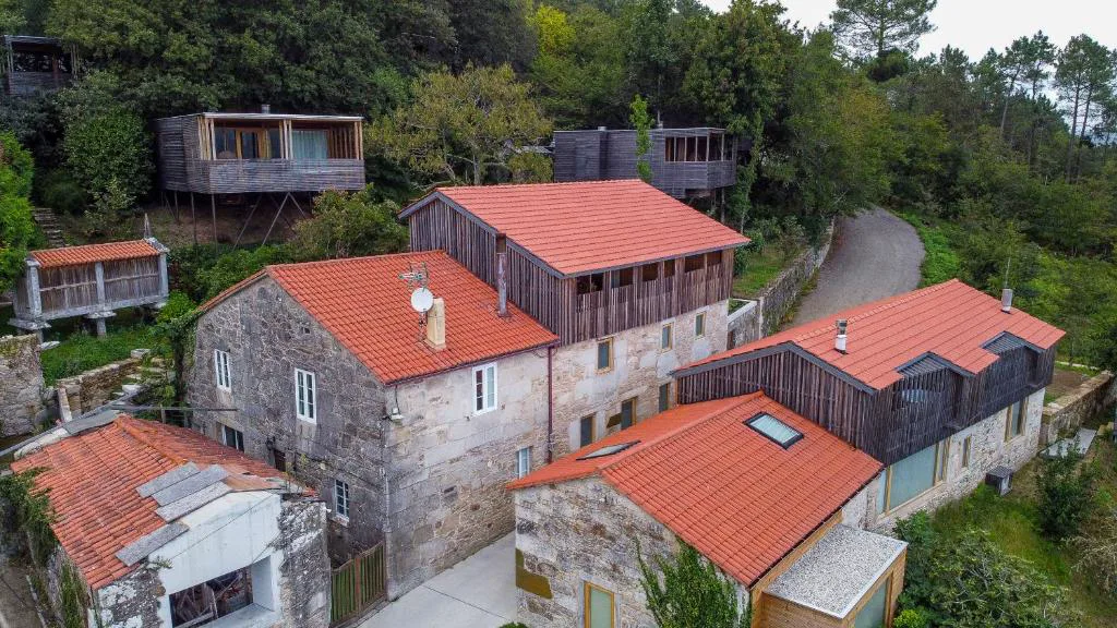 Vista aérea del conjunto de cabañas de madera y hórreos de piedra entre árboles, Cabañas con vista a la ría
