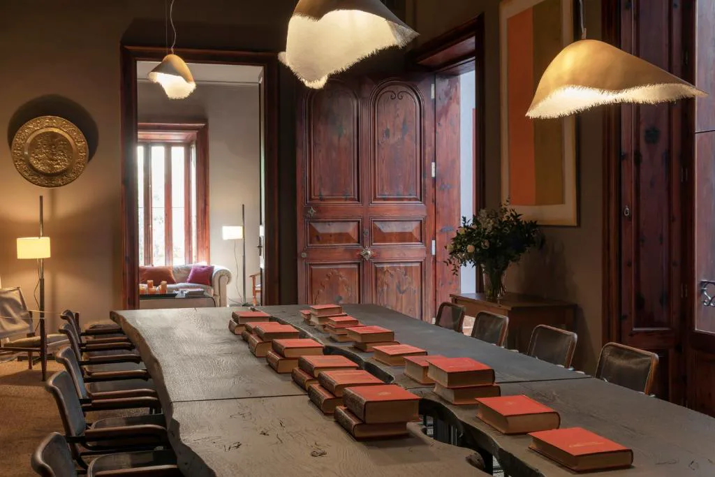 Sala con mesa de biblioteca, libros encuadernados en rojo y puerta antigua de madera tallada, Can Cera Hotel