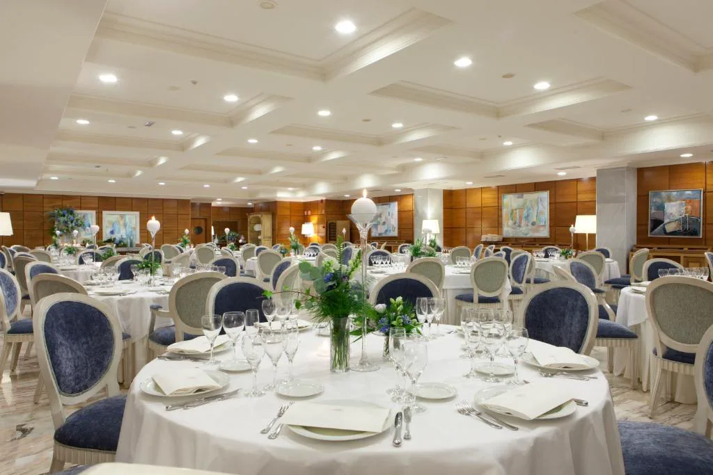 Salón con mesas redondas vestidas de blanco y azul junto a grandes ventanales, Hotel Carlos I Silgar