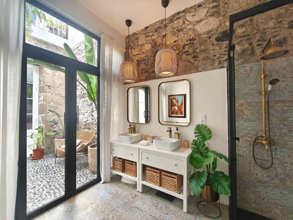 Baño de diseño con doble lavabo, pared de piedra vista y puerta acristalada a un patio con plantas, Casa Bailarina, Agaete, Gran Canaria