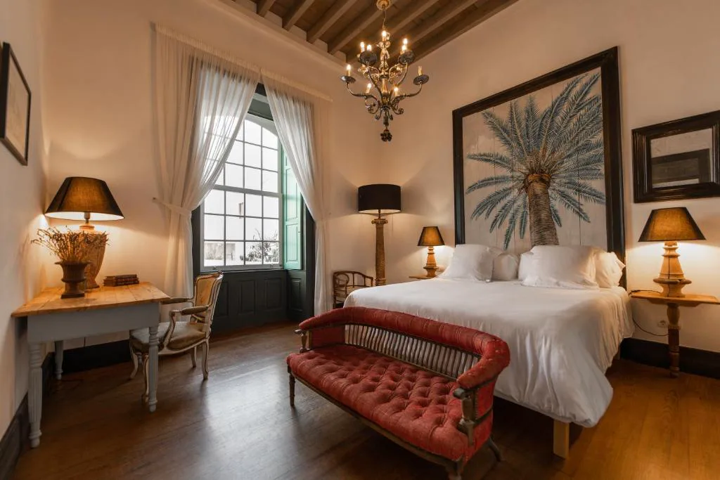 Dormitorio con araña de cristal, cabecero con mural de palmera y sofá de terciopelo rojo, Hotel Emblemático La Casa de los Naranjos
