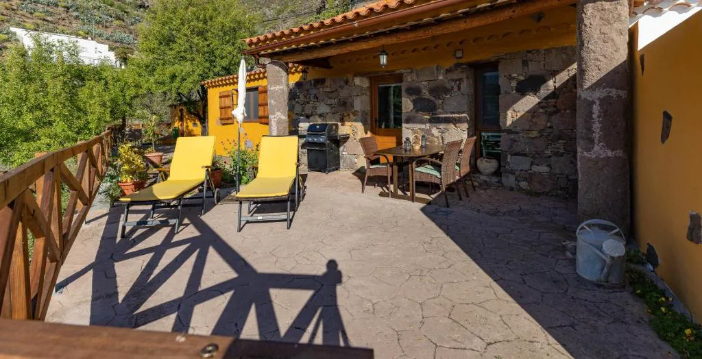 Terraza exterior con tumbonas amarillas, barbacoa y mesa junto a la fachada de piedra y ocre, Casa Rural Bentayga