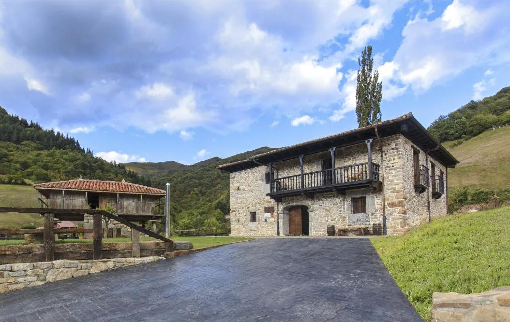 Casa de piedra con balcón de madera junto a un camino, con montañas al fondo