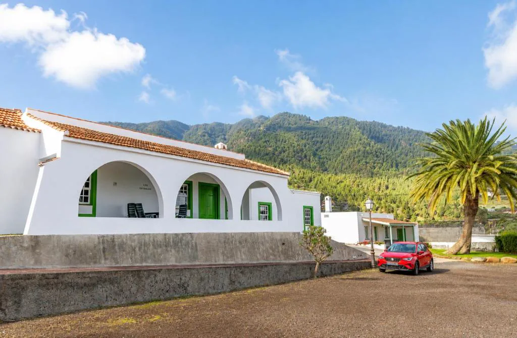 Fachada blanca de arcos y carpintería verde de Casa de Los Establos, con las montañas de La Palma al fondo
