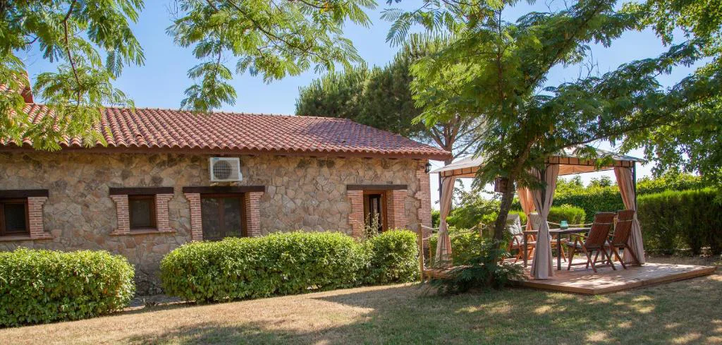 Casita de piedra y teja junto a arbustos y una pérgola