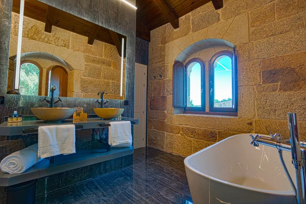 Baño con lavabos de cerámica dorada y bañera exenta junto a ventana de piedra, Hotel As Torres da Hermida