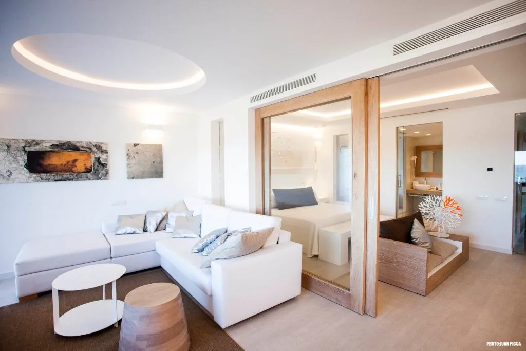 Salón de la suite con sofá blanco y vista a la habitación con ducha acristalada, Hotel Cala Saona & Spa