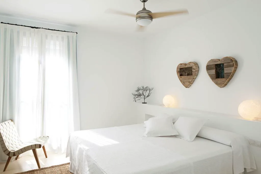 Dormitorio blanco con corazones de madera en la pared y ventilador de techo, Hotel Es Marès