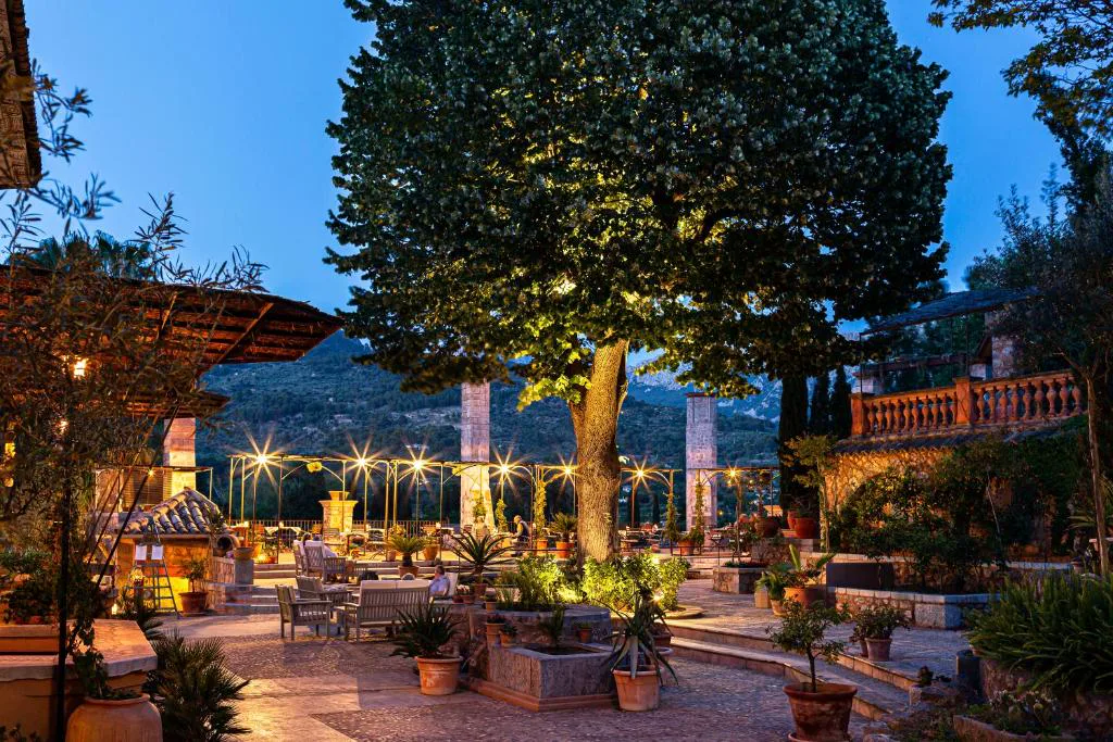 Terraza al atardecer con mesas iluminadas bajo un gran árbol y vistas a la sierra, Hotel Finca Ca N'ai