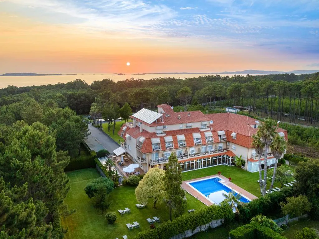 Vista aérea del hotel entre pinares con piscina exterior y el Atlántico al fondo al atardecer, Hotel Spa Atlántico San Vicente do Mar