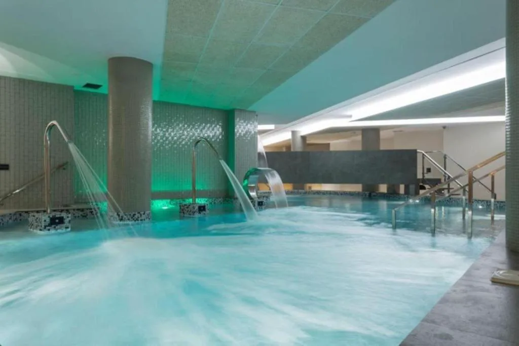Piscina de hidroterapia interior con chorros y cascadas iluminada en tonos verdes, Hotel Spa Odeón
