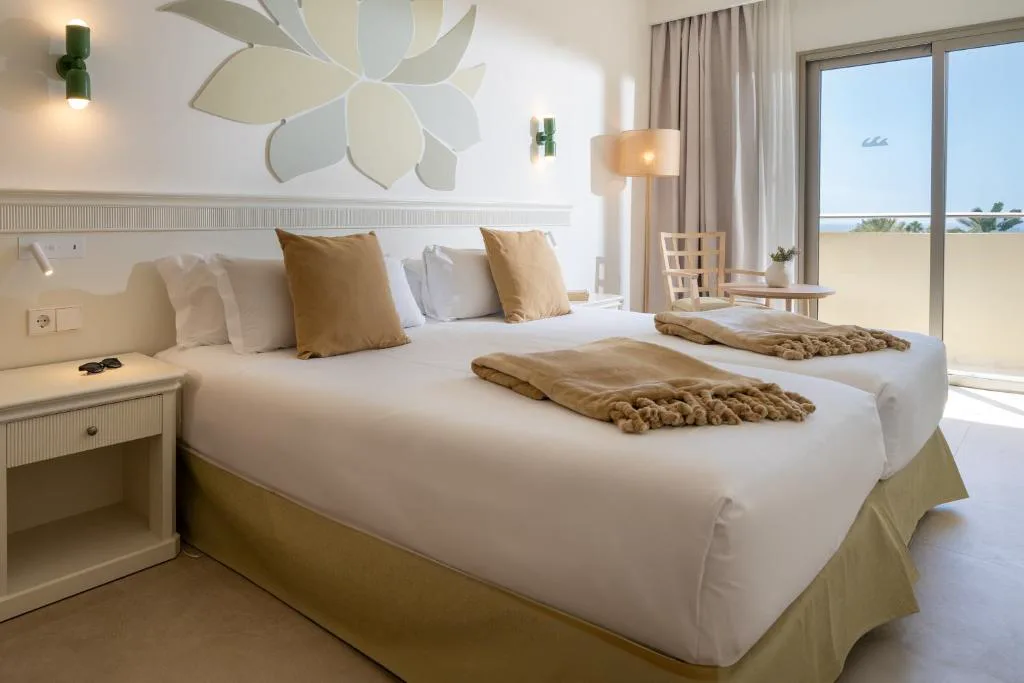 Dormitorio con ropa de cama beige y balcón con vistas a palmeras, Iberostar Waves Gaviotas Park