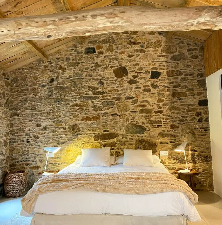 Dormitorio abuhardillado con pared de piedra vista y viga de madera en bruto, Lar do Gado