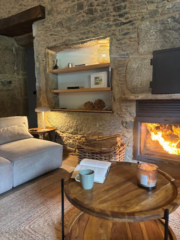 Chimenea de piedra encendida junto a sofá y mesa con libro y vela, Lar do Gado