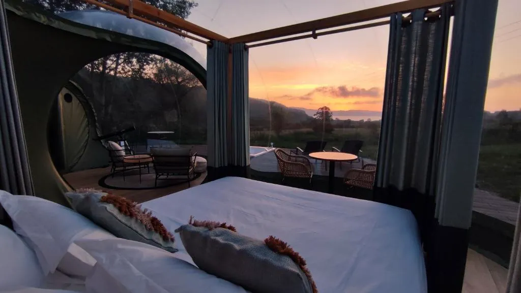 Cama doble bajo la burbuja transparente de Luzada Glamping, con vistas al atardecer sobre el campo en Xove