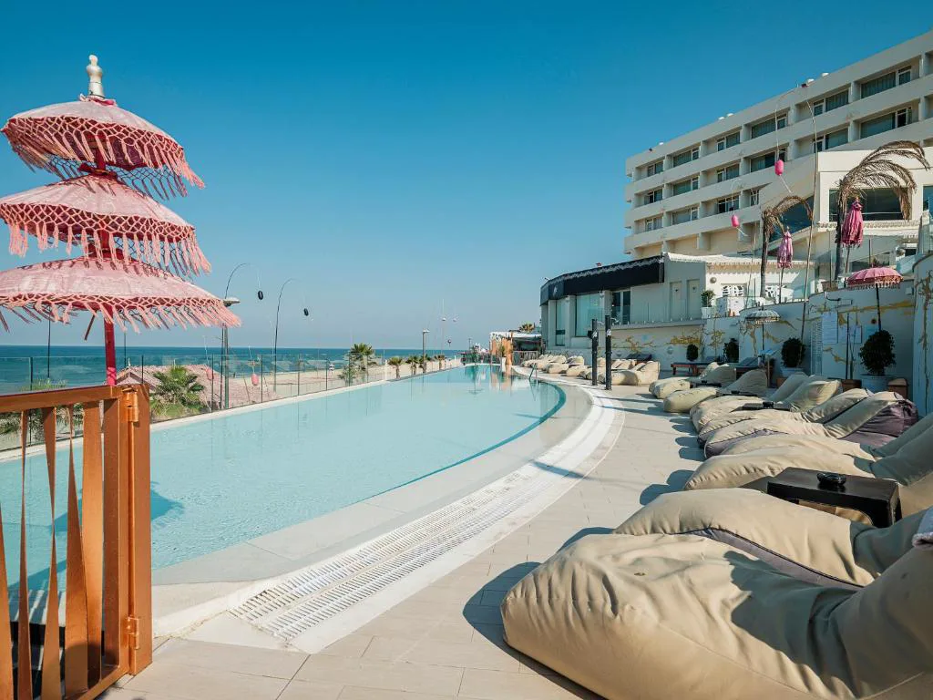 Piscina con sombrilla rosa y pufs frente al hotel