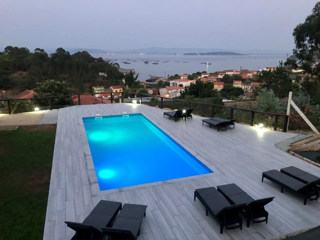 Piscina exterior iluminada al atardecer con la Ría de Arousa y el pueblo al fondo, Cabañas Rías Baixas